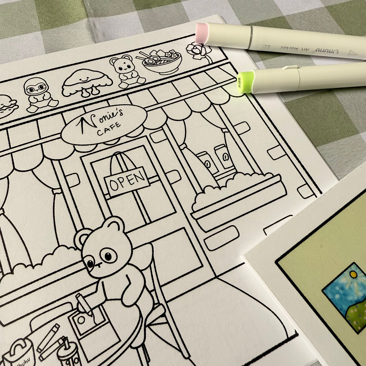 Coloring book page with a teddy bear at a cafe, markers on a checkered tablecloth.