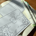 Open notebook with coloring book pages featuring teddy bears and flowers, with markers on a wooden surface.