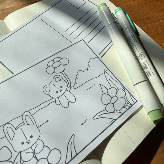 Open notebook with coloring book pages featuring teddy bears and flowers, with markers on a wooden surface.